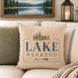 May include: A square beige throw pillow featuring a dark blue and olive green graphic print. The design includes a cabin surrounded by pine trees above the text LAKE WEEKEND and a small oar icon, all enclosed within a thin rectangular border.