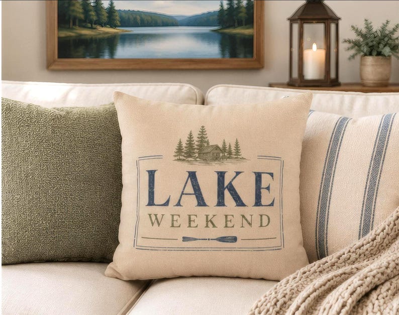 May include: A square beige throw pillow featuring a dark blue and olive green graphic print. The design includes a cabin surrounded by pine trees above the text LAKE WEEKEND and a small oar icon, all enclosed within a thin rectangular border.
