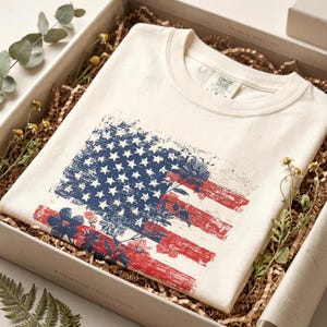 May include: Cream-coloured t-shirt with a distressed American flag design, featuring blue stars, red stripes, and floral accents. The shirt is folded in a gift box with decorative paper and sprigs of greenery.