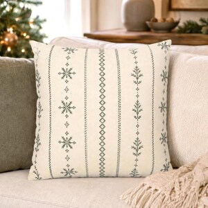 May include: A square throw pillow with an off-white base featuring vertical stripes of dark green embroidered patterns. The design includes repeating snowflake motifs, geometric diamond chains, and stylized pine tree branch silhouettes.