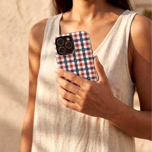 May include: A smartphone case with a red, white, and blue gingham pattern. The case has a black border and a camera cutout. The phone case is held by a person wearing a light-colored sleeveless top.
