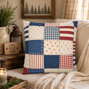May include: Square patchwork pillow with a patriotic theme. The pillow features various patterns, including blue and white stars, red and white stripes, and blue gingham. The pillow is on a beige surface.