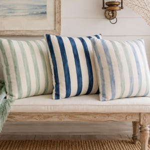 May include: Three square throw pillows featuring vertical watercolor-style stripes. The left pillow has sage green stripes, the center pillow has deep navy blue stripes, and the right pillow has light blue and green stripes on a white background.