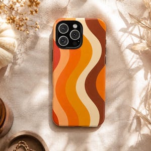 May include: A rectangular smartphone case featuring a vertical wavy pattern in shades of dark brown, cream, bright orange, and muted yellow. The case has a black camera cutout area at the top left corner, designed to fit a multi-lens camera system.