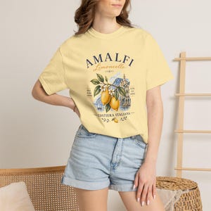 May include: A light yellow t-shirt featuring the text "AMALFI Limoncello" above a graphic of lemons, a coastal scene, and the words "Costiera Italiana." The shirt has short sleeves and a crew neck.