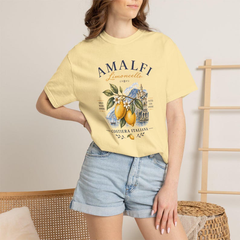 May include: A light yellow t-shirt featuring the text "AMALFI Limoncello" above a graphic of lemons, a coastal scene, and the words "Costiera Italiana." The shirt has short sleeves and a crew neck.