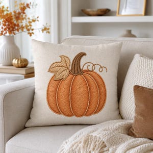 May include: A square throw pillow with a light beige linen-like fabric cover. The center features an embroidered design of a large orange pumpkin with a brown stem, a single tan leaf, and a small curly vine, all rendered in thick thread.