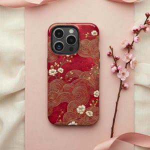 May include: A red phone case with a gold swirling pattern and white floral accents. The case has a wood-grain top and a triple-lens camera cutout. A sprig of pink flowers and a pink ribbon are in the background.