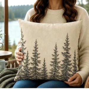 May include: A square throw pillow featuring a cream-colored fabric base with a dark green, detailed print of a forest of pine trees across the bottom half. The design shows multiple evergreen trees of varying heights, creating a nature-inspired scene.