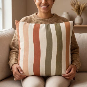 May include: A square throw pillow featuring a vertical striped pattern in shades of cream, light peach, terracotta orange, and muted olive green. The fabric has a smooth, matte texture and the stripes are of varying widths across the front.