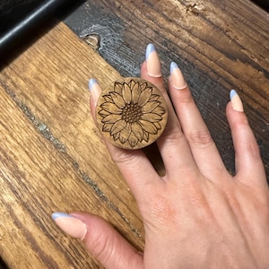 May include: A circular wooden phone grip featuring a laser-engraved sunflower design with detailed petals and a textured center. The natural light brown wood grain is visible across the surface of the round accessory, which is designed to attach to a device.