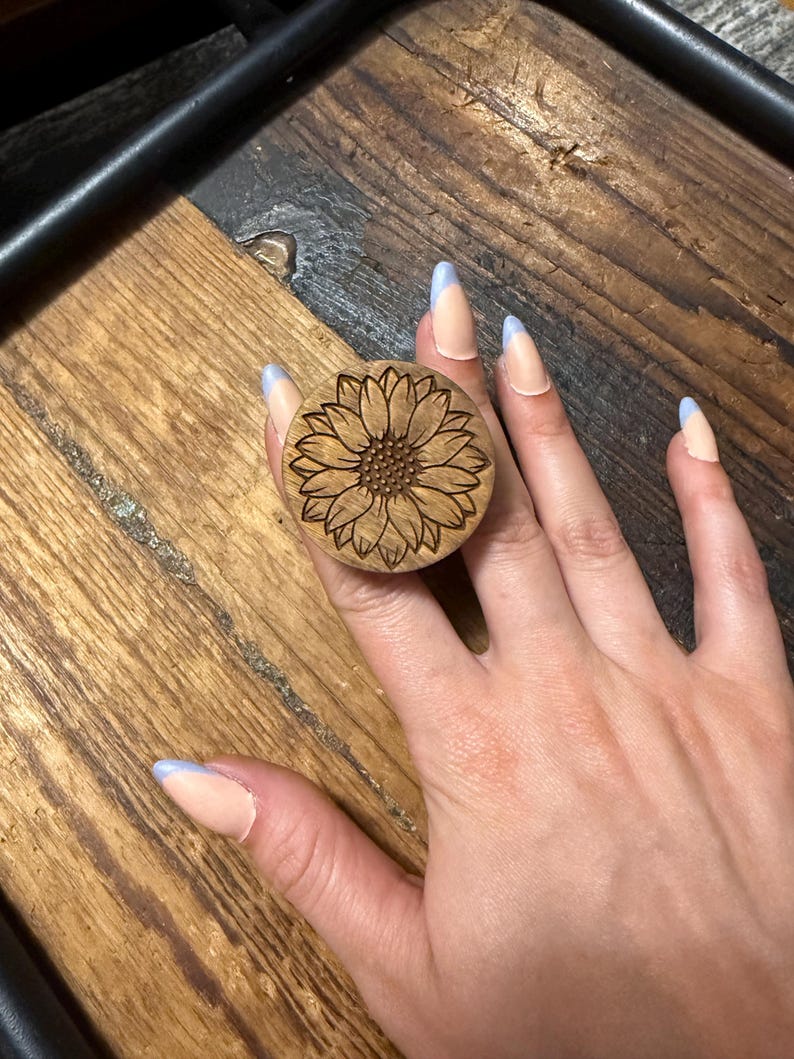 May include: A circular wooden phone grip featuring a laser-engraved sunflower design with detailed petals and a textured center. The natural light brown wood grain is visible across the surface of the round accessory, which is designed to attach to a device.