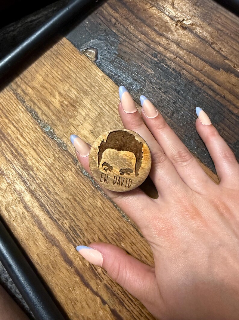May include: A circular wooden phone grip featuring a laser-engraved illustration of a person's eyes and hair, with the text "EW, DAVID" etched clearly below the image. The natural wood grain is visible on the surface of the round accessory.