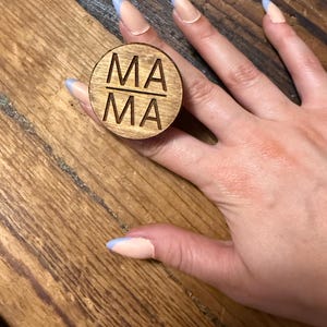 May include: A circular wooden ring featuring the word MA separated by a horizontal line, with the text MA engraved into the surface. The wood has a natural brown grain finish, and the design is centered on the flat, circular top of the ring.