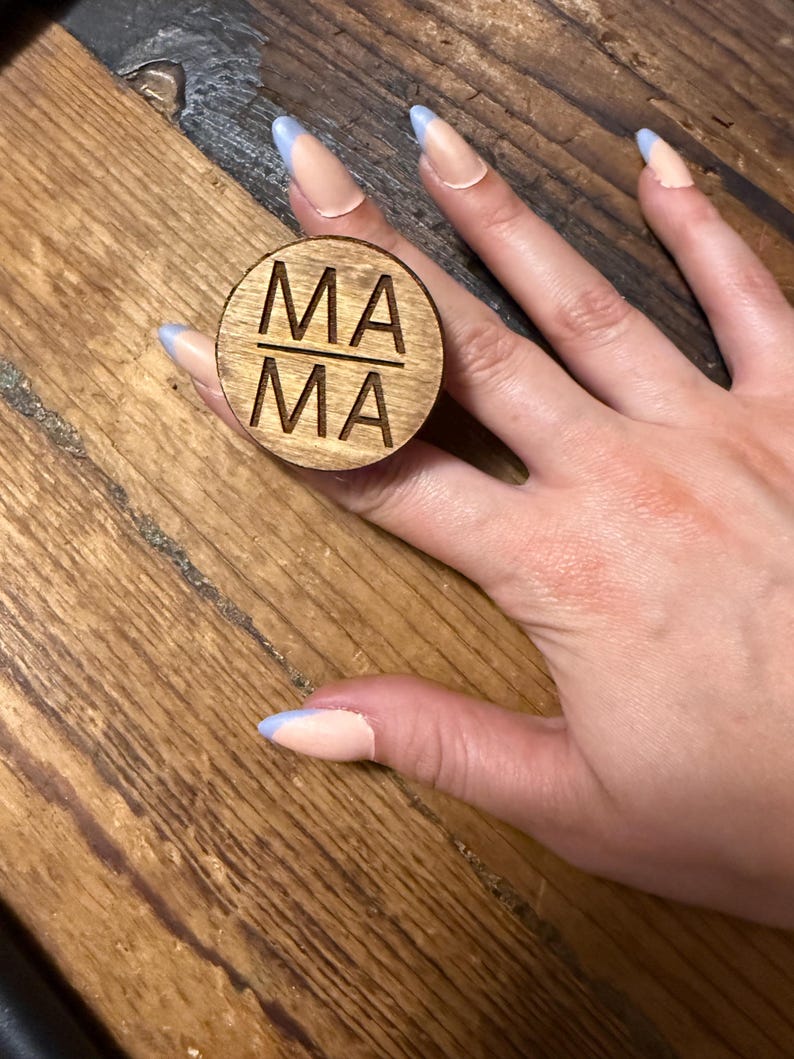 Op de afbeelding: Een ronde houten ring met het woord MA gescheiden door een horizontale lijn, waarbij de tekst MA in het oppervlak is gegraveerd. Het hout heeft een natuurlijke bruine nerf en het ontwerp is gecentreerd op de platte, ronde bovenkant.