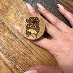 May include: A circular wooden phone grip featuring a laser-engraved design of a person with a messy bun hairstyle, a patterned headband, and sunglasses decorated with a star and stripe motif, creating a patriotic aesthetic on the natural wood surface.