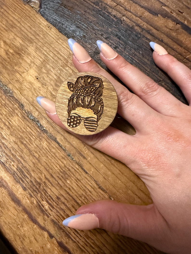 May include: A circular wooden phone grip featuring a laser-engraved design of a person with a messy bun hairstyle, a patterned headband, and sunglasses decorated with a star and stripe motif, creating a patriotic aesthetic on the natural wood surface.