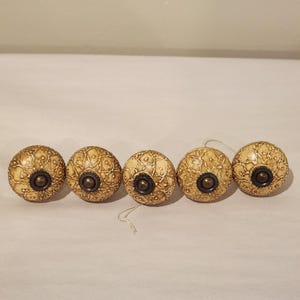 Puede incluir: Cinco pomos redondos para armarios con un patrón floral agrietado en color crema y bronceado. Cada pomo tiene una pieza central de metal en tono bronce oscuro con una pequeña protuberancia circular. Los pomos están en fila horizontal.