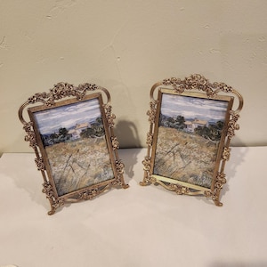 May include: Two identical rectangular picture frames with an ornate gold-toned metal finish. The frames feature intricate rose and floral detailing along the edges and stand on small feet. Each frame holds a print of a landscape painting.