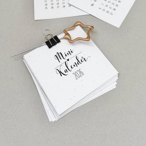May include: A stack of small white calendars with the text "Mini Kalender 2026" in black script. A gold star-shaped paper clip holds the calendar pages together. Another calendar page is visible in the background.