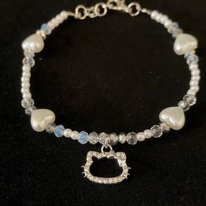 Könnte beinhalten: Ein Armband mit weißen Kunstperlen, klaren und irisierenden Perlen und herzförmigen Akzenten. Ein silberfarbener Anhänger in Form eines Katzenkopfes, besetzt mit kleinen, klaren Kristallen, hängt in der Mitte.