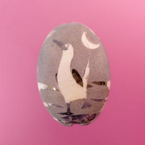 May include: Oval-shaped decorative stone with a painted design of a white bird with a black beak and wing, standing on a gray surface. A white crescent moon is in the upper right corner. The background is a solid pink.