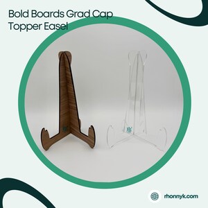 May include: Two easels, one wood-toned and one clear acrylic, are displayed. The wooden easel has a natural grain, while the acrylic one is transparent. Both have a three-legged base and are designed to hold a graduation cap topper. The text "Bold Boards Grad Cap Topper Easel" is visible.