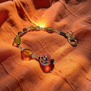 K&ouml;nnte beinhalten: Ein Armband mit Bernstein- und klaren Glasperlen, einem goldenen Verschluss und einer k&uuml;rbisf&ouml;rmigen Perle. Das Armband wird auf orangefarbenem Stoff pr&auml;sentiert. Die Perlen sind auf einer silbernen Kette aufgereiht.