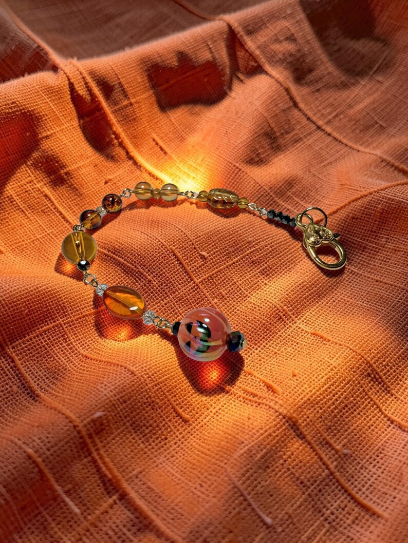 K&ouml;nnte beinhalten: Ein Armband mit Bernstein- und klaren Glasperlen, einem goldenen Verschluss und einer k&uuml;rbisf&ouml;rmigen Perle. Das Armband wird auf orangefarbenem Stoff pr&auml;sentiert. Die Perlen sind auf einer silbernen Kette aufgereiht.