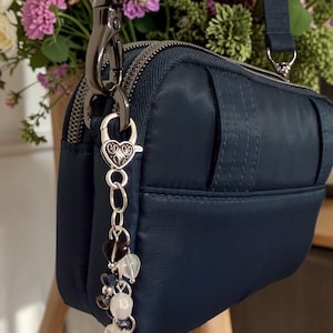 May include: Navy blue crossbody bag with a silver zipper and hardware. A decorative charm with heart-shaped beads and crystals hangs from the zipper. The bag has a matching navy blue strap and is set against a backdrop of colorful flowers.
