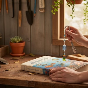 May include: A decorative beaded charm with blue and green accents is held above a book titled "Ocean Life: Comprehensive Guide to Fish." The book is on a wooden surface with gardening tools and potted plants in the background.