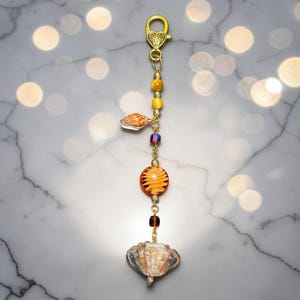 May include: A gold-toned keychain with a lobster clasp, featuring a decorative strand of beads and charms. The strand includes yellow and orange beads, a seashell charm, and a large, round orange glass bead. The bottom charm is a seashell.