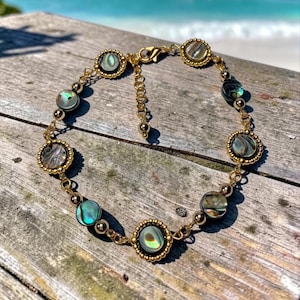 May include: A gold-toned chain bracelet featuring circular abalone shell inlays with iridescent green and blue hues. The bracelet includes small gold-toned metal beads between the shell settings and a lobster claw clasp with an adjustable chain link.