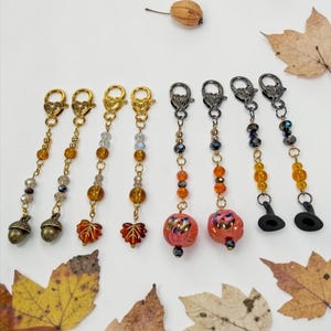 May include: A collection of eight decorative zipper pulls featuring gold and gunmetal lobster clasps. The charms include metal acorns, translucent maple leaves, orange pumpkin beads, and black witch hat charms accented with glass beads.