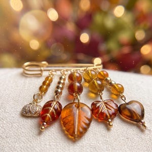 May include: A gold-toned safety pin brooch featuring five dangling charms. The charms include a small crystal-encrusted pumpkin, three amber-colored glass leaves in varying shapes, and a heart-shaped amber glass bead, all accented with gold beads.