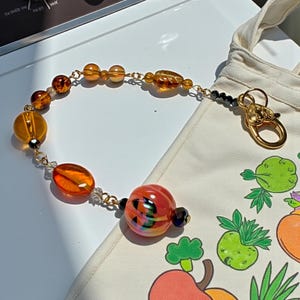 K&ouml;nnte beinhalten: Ein Armband mit einer goldenen Kette und einer Vielzahl von bernsteinfarbenen und orangefarbenen Glasperlen. Das Armband liegt neben einer wei&szlig;en Tragetasche mit einem bunten Fruchtdesign und einem goldenen Verschluss.