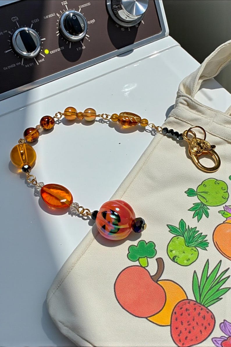 K&ouml;nnte beinhalten: Ein Armband mit einer goldenen Kette und einer Vielzahl von bernsteinfarbenen und orangefarbenen Glasperlen. Das Armband liegt neben einer wei&szlig;en Tragetasche mit einem bunten Fruchtdesign und einem goldenen Verschluss.