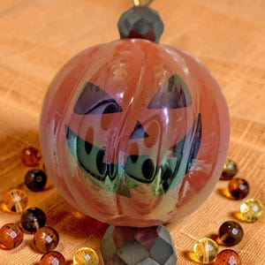 K&ouml;nnte beinhalten: Ein Halloween-Ornament mit einem l&auml;chelnden K&uuml;rbisgesicht mit schwarzen ausgeschnittenen Augen und Mund. Der orangefarbene K&uuml;rbis ist mit schillernden gr&uuml;nen und violetten Farbt&ouml;nen akzentuiert. Das Ornament ist von kleinen, bernsteinfarbenen Perlen umgeben.