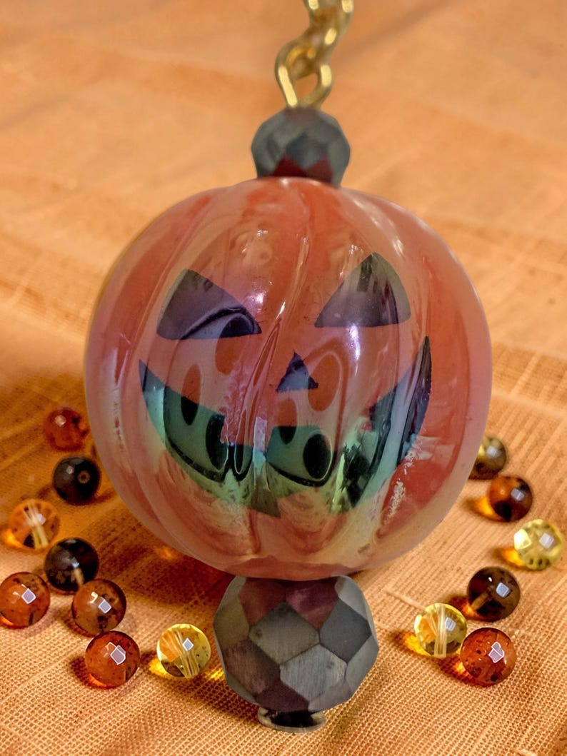 K&ouml;nnte beinhalten: Ein Halloween-Ornament mit einem l&auml;chelnden K&uuml;rbisgesicht mit schwarzen ausgeschnittenen Augen und Mund. Der orangefarbene K&uuml;rbis ist mit schillernden gr&uuml;nen und violetten Farbt&ouml;nen akzentuiert. Das Ornament ist von kleinen, bernsteinfarbenen Perlen umgeben.