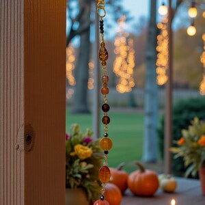 K&ouml;nnte beinhalten: Ein dekoratives H&auml;ngeornament mit einem goldenen Verschluss und einer Kette aus bunten Perlen in Braun-, Orange- und Gelbt&ouml;nen. Das Ornament h&auml;ngt vor einem verschwommenen Hintergrund aus K&uuml;rbissen, Blumen und Lichterketten und schafft eine herbstliche Szene.