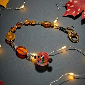 K&ouml;nnte beinhalten: Ein Armband mit bernsteinfarbenen und orangefarbenen Glasperlen in verschiedenen Formen, darunter runde und ovale. Das Armband hat einen goldfarbenen Verschluss und schwarze Perlen. Herbstbl&auml;tter und Lichterketten umgeben das Armband.