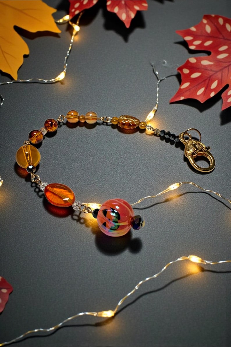 K&ouml;nnte beinhalten: Ein Armband mit bernsteinfarbenen und orangefarbenen Glasperlen in verschiedenen Formen, darunter runde und ovale. Das Armband hat einen goldfarbenen Verschluss und schwarze Perlen. Herbstbl&auml;tter und Lichterketten umgeben das Armband.