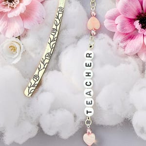 May include: A gold-colored metal bookmark with a floral design, paired with a beaded charm spelling "TEACHER". The charm includes pink heart-shaped beads and a decorative pink glass orb. The background features pink and white flowers.