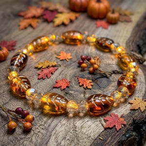 May include: A circular garland featuring amber-colored glass beads shaped like leaves with etched veins, interspersed with clear faceted glass beads that glow with warm internal lights, arranged in a ring on a textured wooden surface.