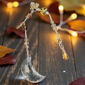 May include: A decorative hanging ornament featuring a clear, faceted crystal crescent moon suspended from a silver-toned chain. The chain is adorned with various amber, gold, and clear glass beads, including a small heart-shaped bead and a teardrop pendant.