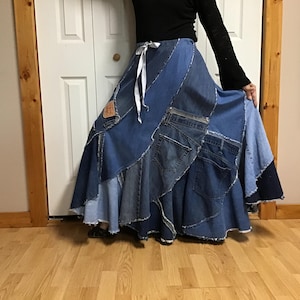 May include: A floor-length skirt constructed from various shades of blue denim fabric patches. The skirt features a white ribbon drawstring at the waist, visible back pockets from jeans, and frayed edges along the seams of the patchwork design.