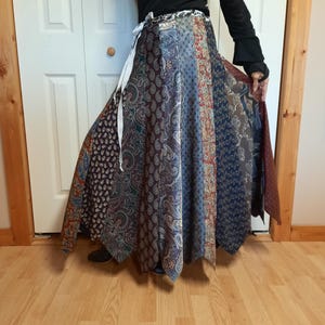 May include: A long, flowing skirt made from repurposed neckties. The skirt is a patchwork of different patterns and colors, including paisley, stripes, and solids. The skirt is tied at the waist with a white ribbon.