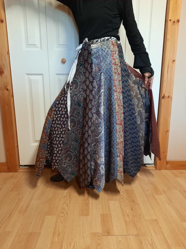 May include: A long, flowing skirt made from repurposed neckties. The skirt is a patchwork of different patterns and colors, including paisley, stripes, and solids. The skirt is tied at the waist with a white ribbon.