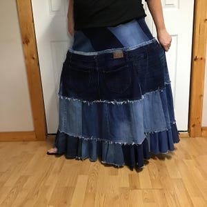 May include: A floor-length tiered skirt constructed from various shades of blue denim fabric. The design features multiple panels with raw, frayed edges and includes a prominent dark blue denim waistband section with visible back pockets and a Lee label.