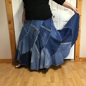 May include: A floor-length patchwork skirt constructed from various shades of blue denim fabric. The skirt features multiple panels with raw, frayed edges and visible back pockets from repurposed jeans, creating a flared, voluminous silhouette.